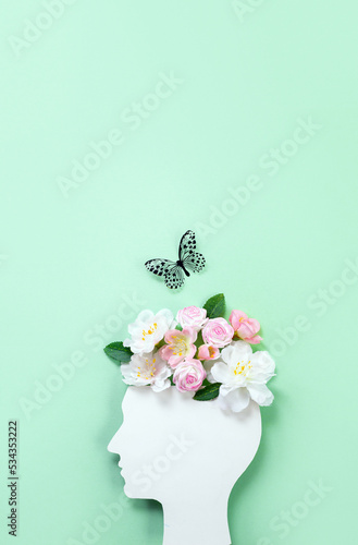 Cardboard silhouette of human head decorated with flowers and butterfly on mint background. World mental health day concept. Vertical banner. Copy space