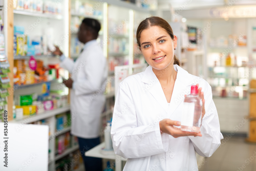 Portrait of a smiling young female pharmacist in the sales hall of a pharmacy, demonstrating recently received goods for ..sale in hands