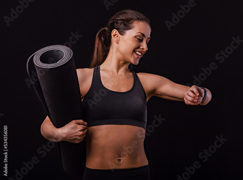 Happy smiling fit healthy fitness woman in bra top clothing holding black mat for yoga and looking on watches what time is it now on black background with empty copy space. Closeup