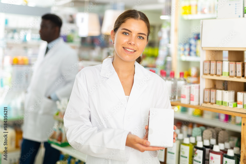Positive young female pharmacist standing in the sales hall of a pharmacy shows recently received packs of vitamins for sale ..in her hands