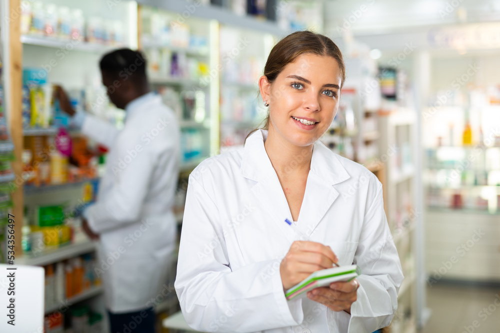 Female pharmacist standing in salesroom of drugstore with notebook in hands. Her colleague working behind.