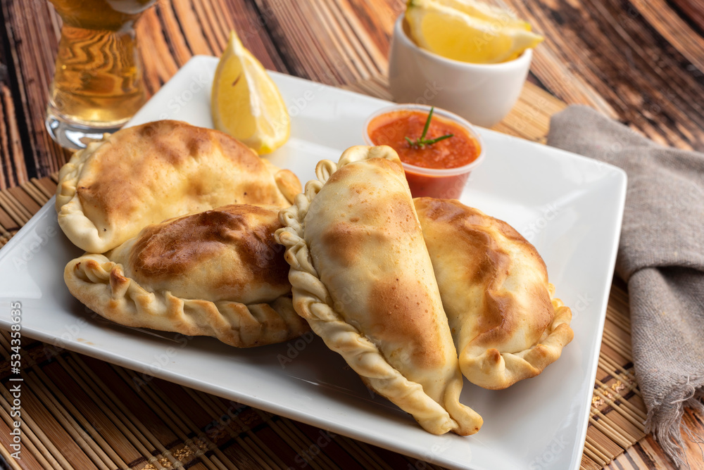 Argentinian homemade empanadas on square white plate red sauce and lemon slices on wooden table Argentinian homemade empanadas on square white plate red sauce and lemon slices on wooden table