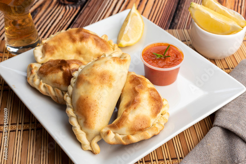 Argentinian homemade empanadas on square white plate red sauce and lemon slices on wooden table