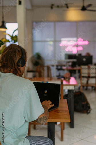 Young man in casual wear with headphones works on a laptop in a coworking space in Bali. Distant work.