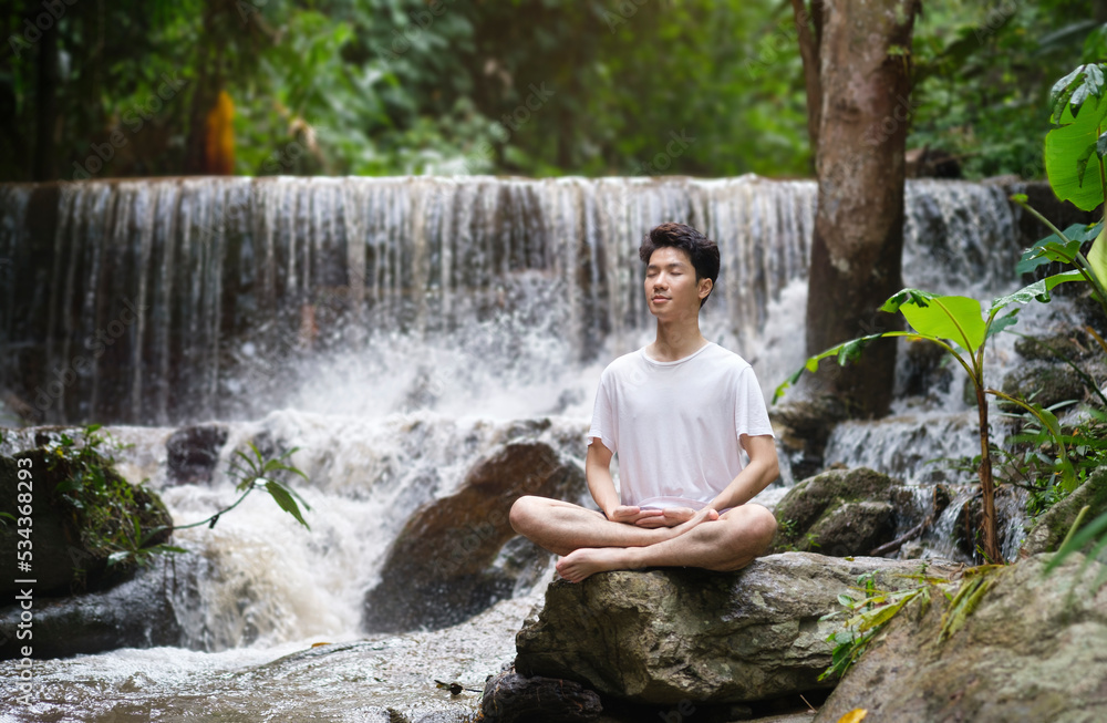 Peaceful young man practicing yoga meditation near waterfall in forest. Concept of calm and meditation.