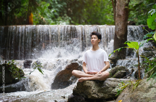 Peaceful young man practicing yoga meditation near waterfall in forest. Concept of calm and meditation.