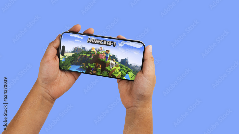 Fototapeta premium Child holding a smartphone iPhone 14 Pro with Minecraft mobile game app on the screen. Blue background. Rio de Janeiro, RJ, Brazil. September 2022