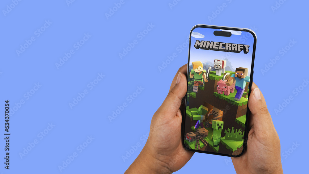 Fototapeta premium Child holding a smartphone iPhone 14 Pro with Minecraft mobile game app on the screen. Blue background. Rio de Janeiro, RJ, Brazil. September 2022