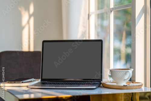 Obraz na płótnie Mockup of laptop computer with empty screen with coffee cup and smartphone on table beside the window of the coffee shop background,Black gray screen w salonie