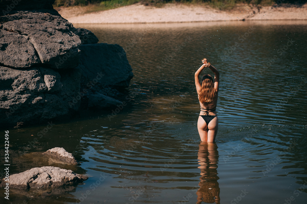 Fototapeta premium A young girl with long blond hair, a slender figure and a beautiful back, in a black swimsuit, stands in a river against the backdrop of rocks.
