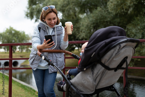 a young mother sipping coffee in headphones walks in the park with a baby stroller