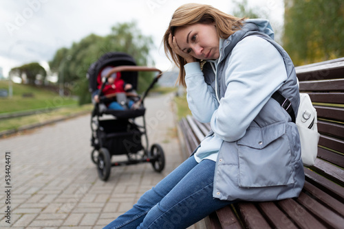 young woman mother of postpartum depression sits with built-in feelings in the park on a bench next to a baby stroller