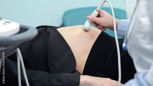 Doctor hand with ultrasonic device examines pregnant businesswoman. Ultrasound technician in white coat scans development and condition of embryo in abdomen