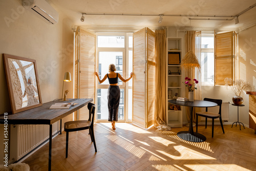 Woman opens window blinds letting the sun inside the room, spending good morning in sunny and cozy apartment in beige tones. Interior view
