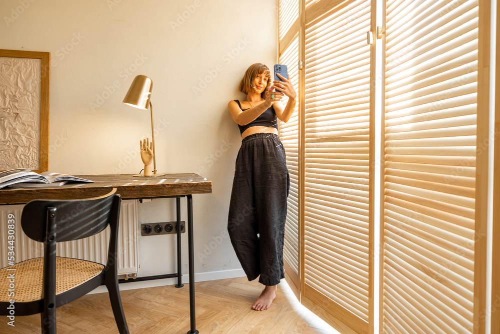 Young stylish woman in black home wear talking online or making photo on smart phone while standing near the window blinds and workplace at home. Concept of blogging and domestic lifestyle