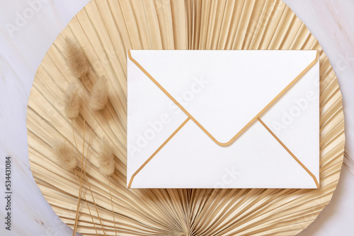 Blank envelope on dry beige palm leaf near hare's tail grass top view,  boho wedding mockup