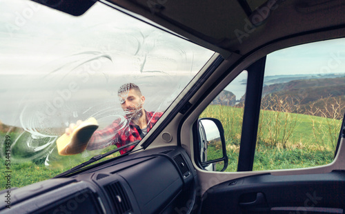 Young man cleaning camper van windshield with sponge outdoor