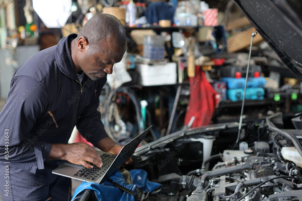 Man technician car mechanic in uniform checking maintenance a car service with computer at repair garage station. Car repair service concept Man technician car mechanic in uniform checking maintenance a car service with computer at repair garage station. Car repair service concept