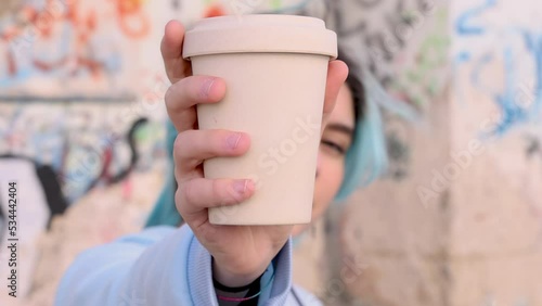 Teenager girl in blue oversize hoodie showing coffee to go cup. Blue haired teen girl staying outdoors near graffiti wall. Mockup, head shot. Adolescence concept