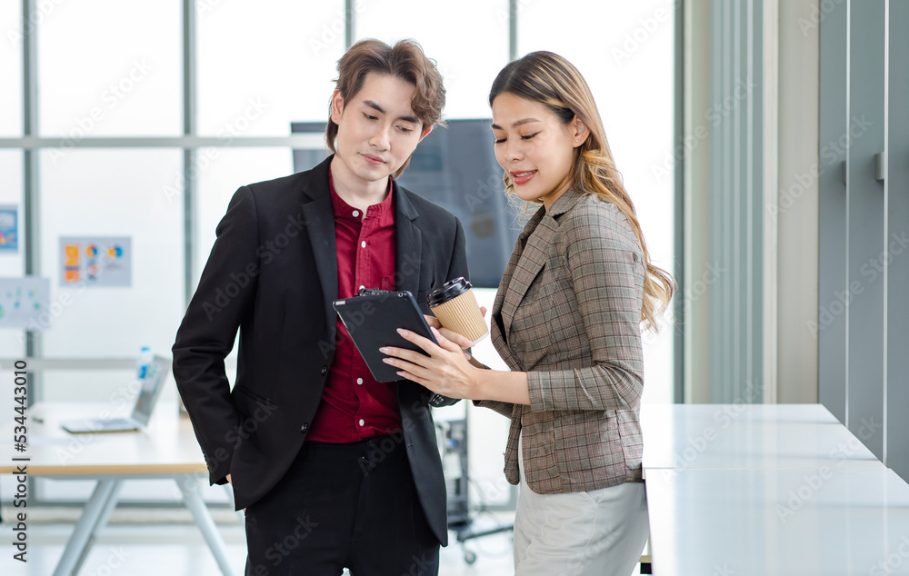 Millennial Asian successful professional female businesswoman in formal suit standing smiling showing data information on touchscreen tablet computer to male businessman colleague in company office