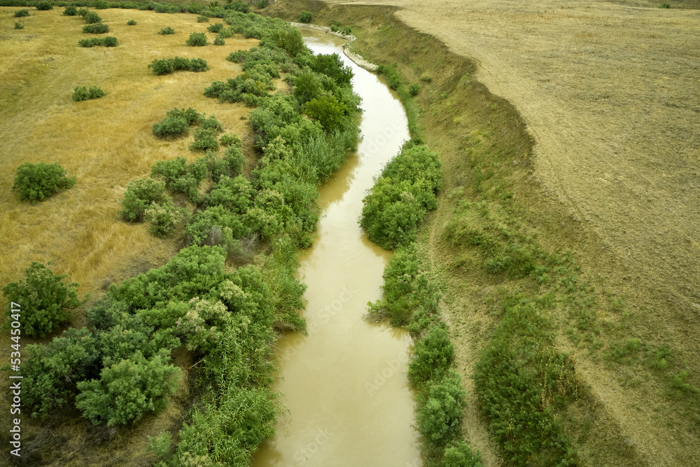 a winding muddy river with overgrown green banks in a sun-scorched steppe rural landscape a winding muddy river with overgrown green banks in a sun-scorched steppe rural landscape
