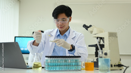 Smiling asian male researcher in white coat conducting experiment with test tubes and microscope slides in research laboratory