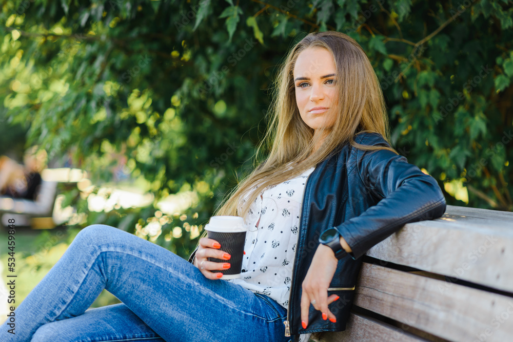 A beautiful fashion young woman sitting on a bench.A girl enjoys coffee from a paper cup.Summer,autumn day.Klaipeda,Lithuania.09-02-2022. A beautiful fashion young woman sitting on a bench.A girl enjoys coffee from a paper cup.Summer,autumn day.Klaipeda,Lithuania.09-02-2022.