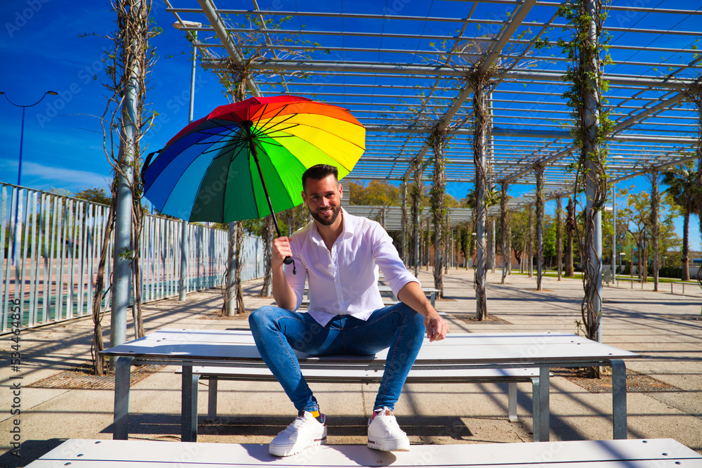 Handsome young blue-eyed gay man is sitting in a park and holding a rainbow-coloured umbrella in his hand. The man is happy. Rights, homosexuality, lesbians.