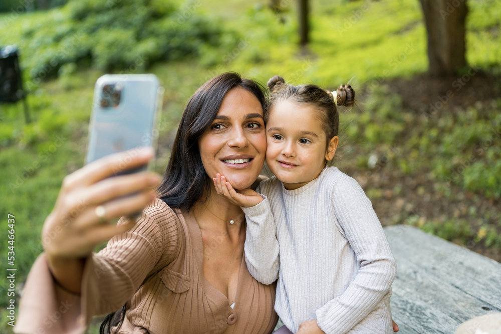 Pregnant white woman and little girl taking selfie together while sitting on bench in park. enjoy beautiful fall day. Pregnant white woman and little girl taking selfie together while sitting on bench in park. enjoy beautiful fall day.