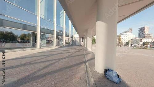 FPV drone view of Modern building of the House of Culture with a glass facade and columns in big city. The copter flies between the columns in zigzags. Autumn sunny day
