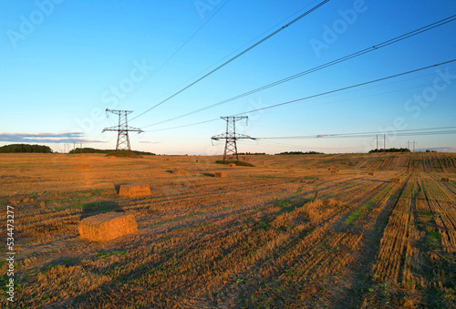 High voltage electricity pylons and transmission power lines in field. Electric tower in field with hay stacks. Power line towers in farmland with hay bales. Hay making on sunset. Electricity pylons.