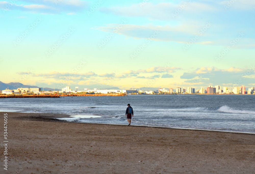 Man walks along the coastline of the sea in autumn season. Young man walks barefoot along the beach near sea at sunset. Person walking on empty beach in winter season. Tourist travels on mediterranean