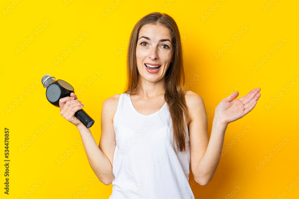 Joyful young woman holding a microphone on a yellow background. Reporter or journalist concept