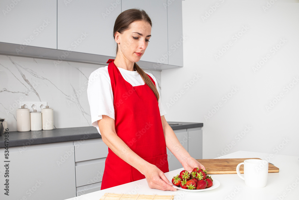 Young woman in red apron holding a plate of strawberries in the kitchen