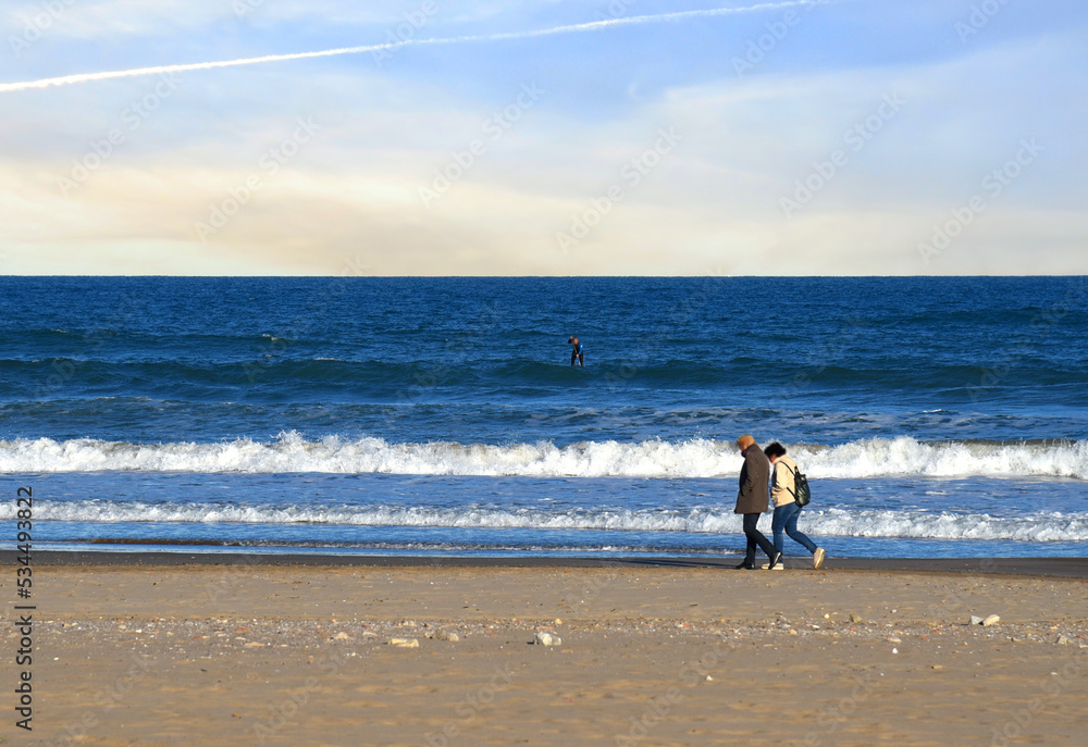People walk along the coastline of the sea in autumn. Man and a woman walk barefoot on beach near sea. Persons walking on empty beach in winter season. Sea beach. Tourist travels in the mediterranean.