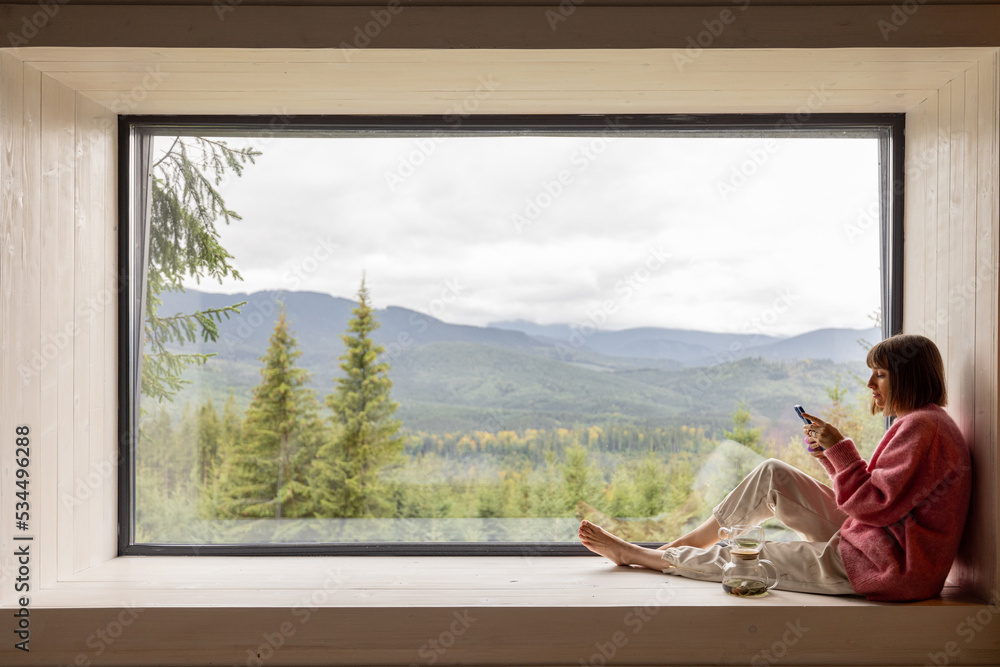 Woman sits with phone on window sill and enjoys scenic view on mountains  while resting in wooden house on nature. Recreation and escaping to nature  Stock Photo