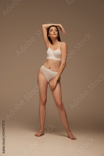Full-length portrait of young beautiful woman posing in white underwear isolated over dark beige studio background. Wellness
