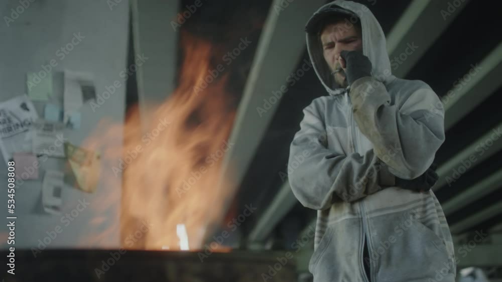 Low angle shot of homeless man in hoodie smoking cigarette and looking pensively at fire burning in barrel while standing in abandoned building Low angle shot of homeless man in hoodie smoking cigarette and looking pensively at fire burning in barrel while standing in abandoned building