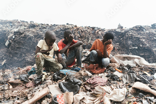 Group of African boys sitting among garbage and plastic waste in an illegal landfill; health risk for street children photo
