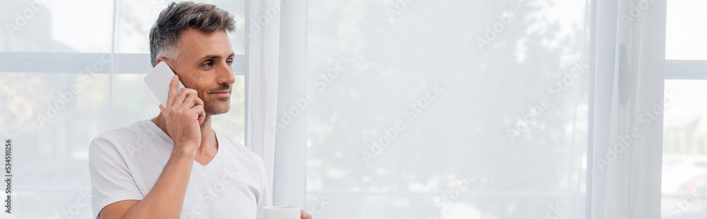 Man in white t-shirt talking on smartphone and holding cup at home, banner. Man in white t-shirt talking on smartphone and holding cup at home, banner.