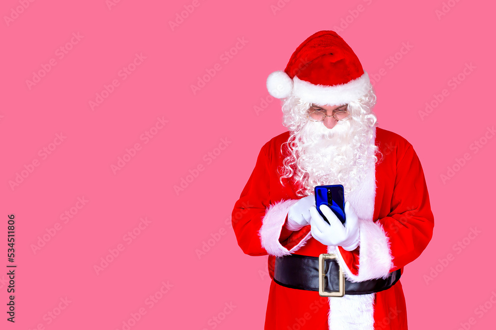 Funny old bearded Santa Claus in a suit holding a mobile phone with a mobile app on a smartphone, shocked by a Christmas advertisement isolated on a pink background. Funny old bearded Santa Claus in a suit holding a mobile phone with a mobile app on a smartphone, shocked by a Christmas advertisement isolated on a pink background.