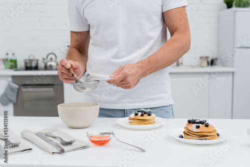 Cropped view of man pouring powdered sugar in sieve near honey and pancakes in kitchen.