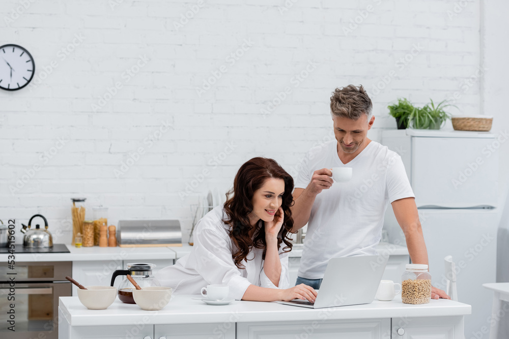 Smiling woman in shirt using laptop near husband with coffee and cereal rings at home.