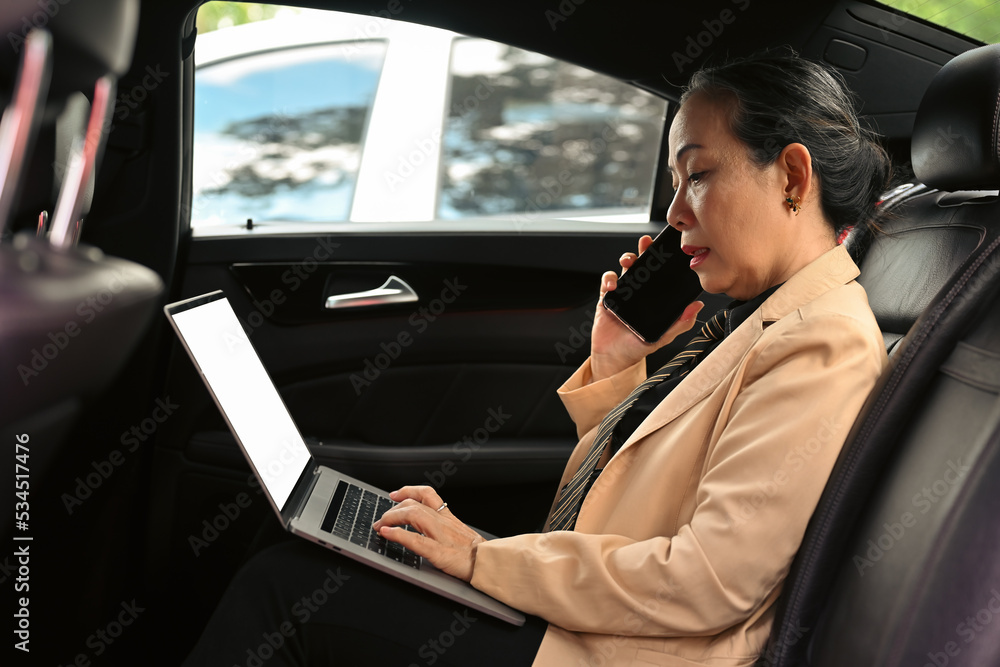 Confident old Asian executive woman in formal clothes concentrating on screen of laptop and interacting with smartphone while sitting in luxurious car. Confident old Asian executive woman in formal clothes concentrating on screen of laptop and interacting with smartphone while sitting in luxurious car.