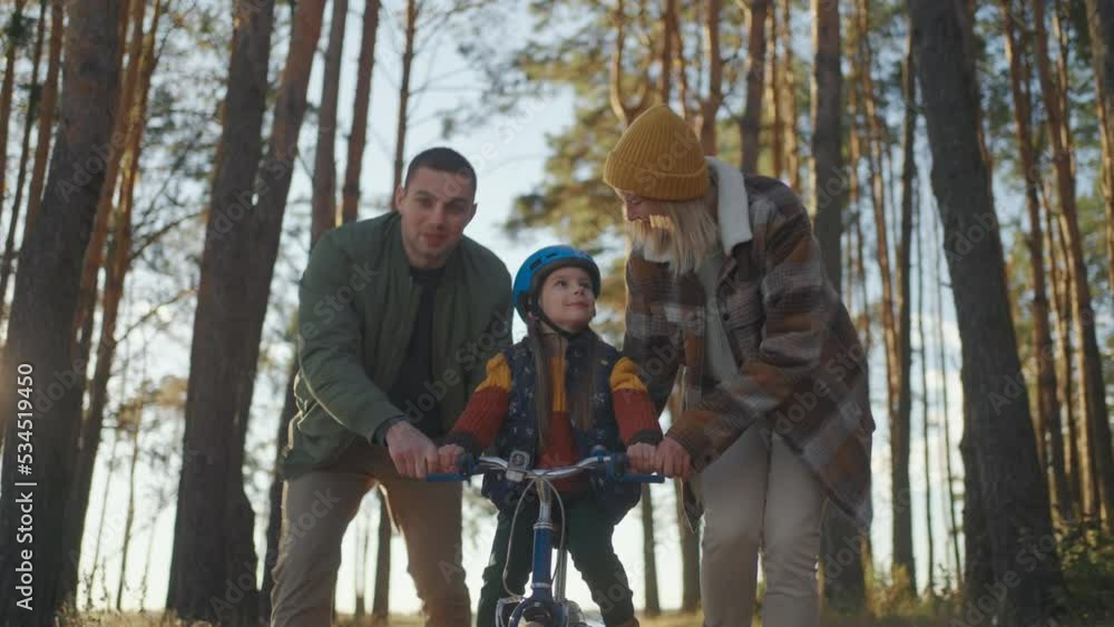 Parents Teaching Daughter To Ride Bike. parents teaching their little girl to driving bike in park. Happy family with excited daughter learning to ride a bike for the first time Parents Teaching Daughter To Ride Bike. parents teaching their little girl to driving bike in park. Happy family with excited daughter learning to ride a bike for the first time