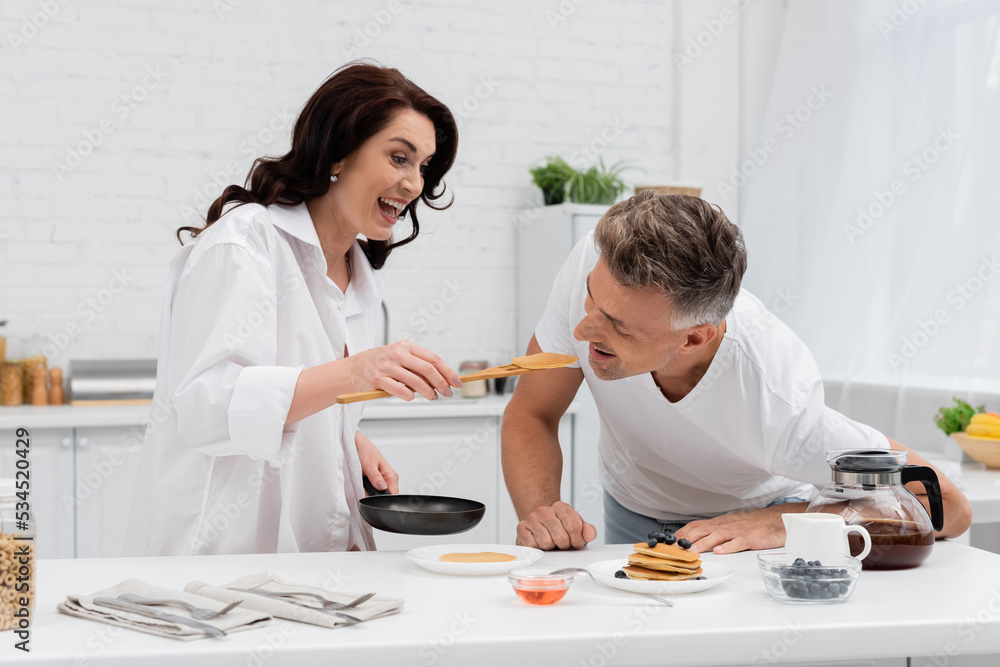 Smiling woman feeding husband with pancake near breakfast in kitchen. Smiling woman feeding husband with pancake near breakfast in kitchen.