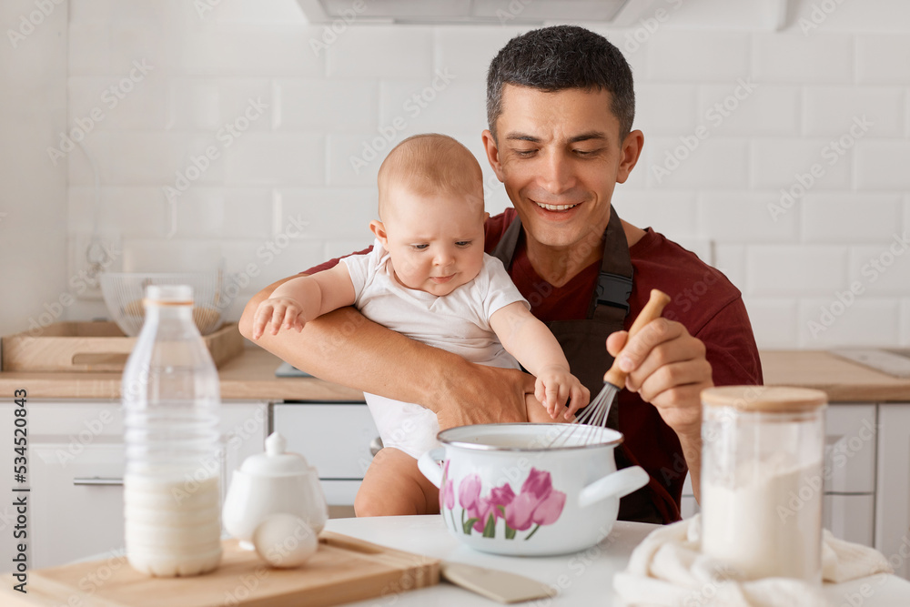 Horizontal shot of satisfied father in apron sitting at table with baby daughter and cooking together in kitchen, mixing dough for homemade pastry, expressing happiness. Horizontal shot of satisfied father in apron sitting at table with baby daughter and cooking together in kitchen, mixing dough for homemade pastry, expressing happiness.