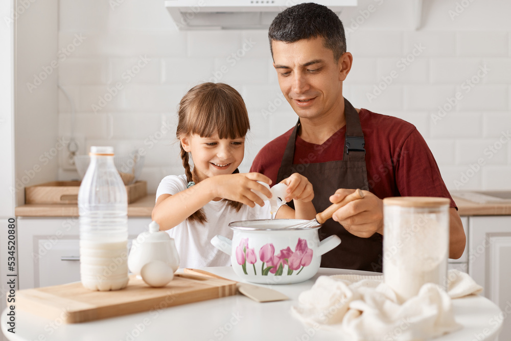 Image of happy family father and kid sitting at table in kitchen, daughter and dad cooking together, adding eggs into dough, preparing delicious surprise for mom. Image of happy family father and kid sitting at table in kitchen, daughter and dad cooking together, adding eggs into dough, preparing delicious surprise for mom.