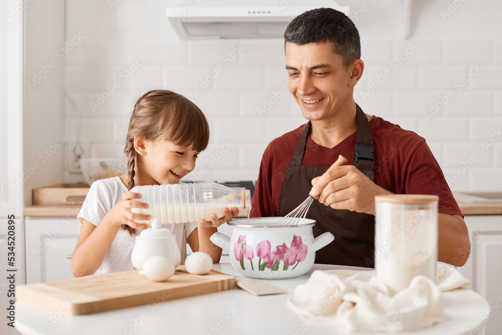 Indoor shot of smiling positive father sitting at table with daughter and cooking together in kitchen, little girl pouring milk into pot and smiling, will making pancakes. Indoor shot of smiling positive father sitting at table with daughter and cooking together in kitchen, little girl pouring milk into pot and smiling, will making pancakes.