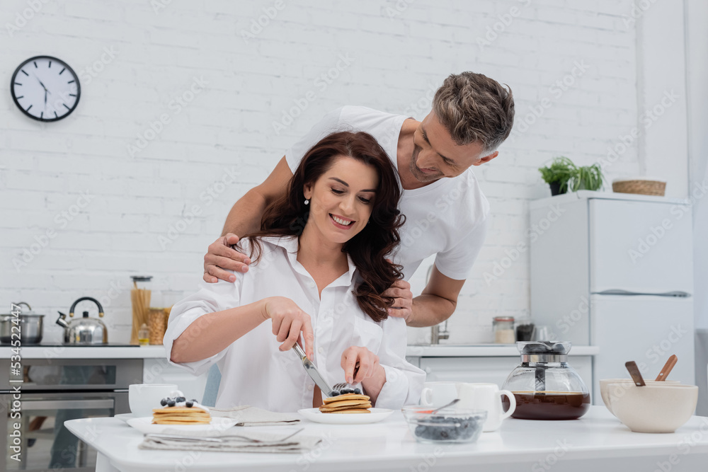 Man hugging smiling wife in shirt cutting pancakes in kitchen.